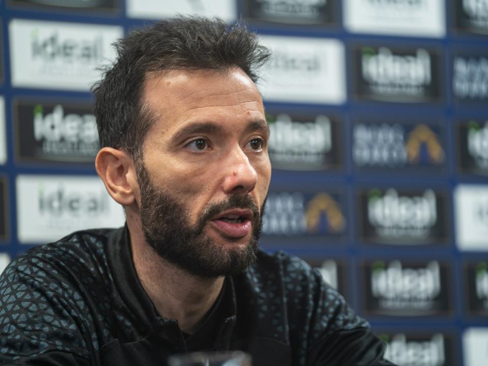 Carlos Corberán speaking at a press conference in front of sponsor boards