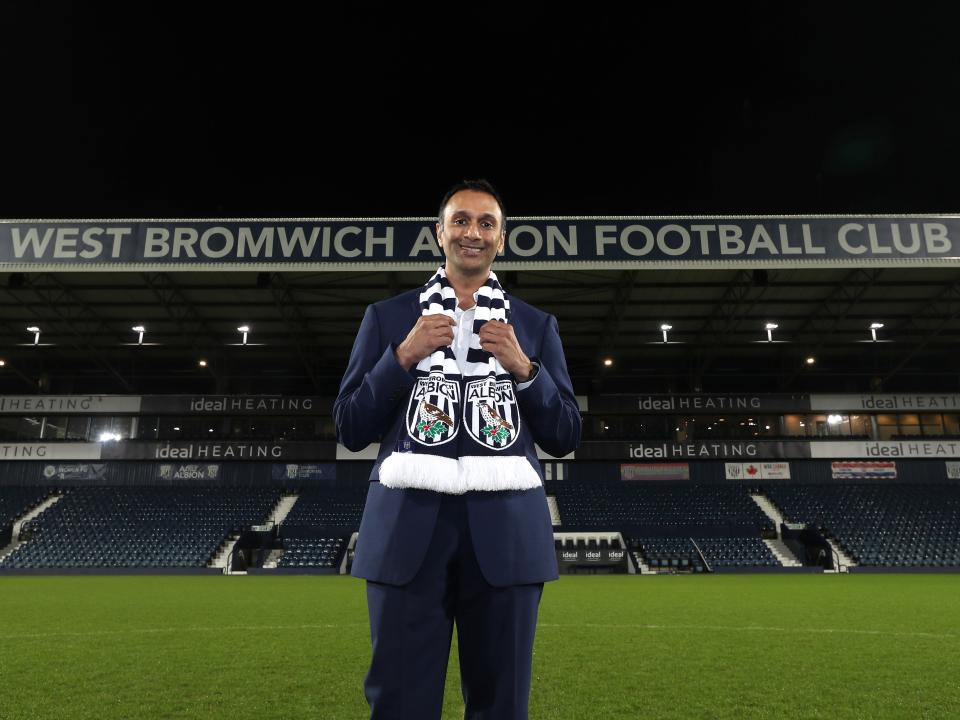 Shilen Patel stood on the pitch at The Hawthorns with the West Stand behind him and an Albion scarf wrapped around his neck