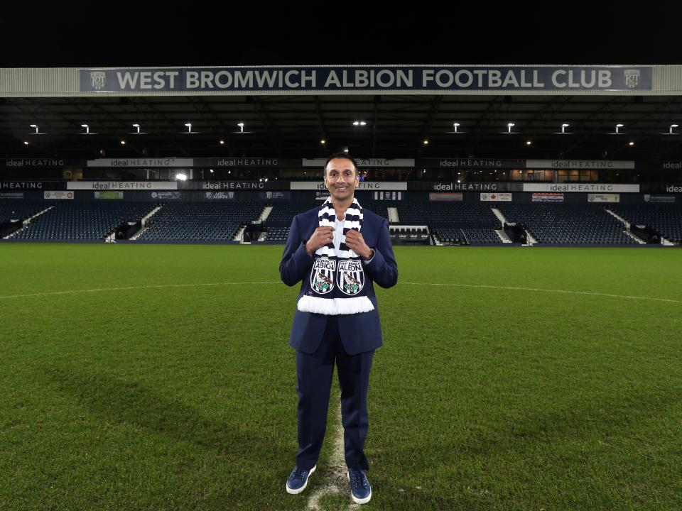 Shilen Patel stood on the pitch at The Hawthorns smiling at camera with the West Stand in the background and an Albion scarf wrapped round his neck