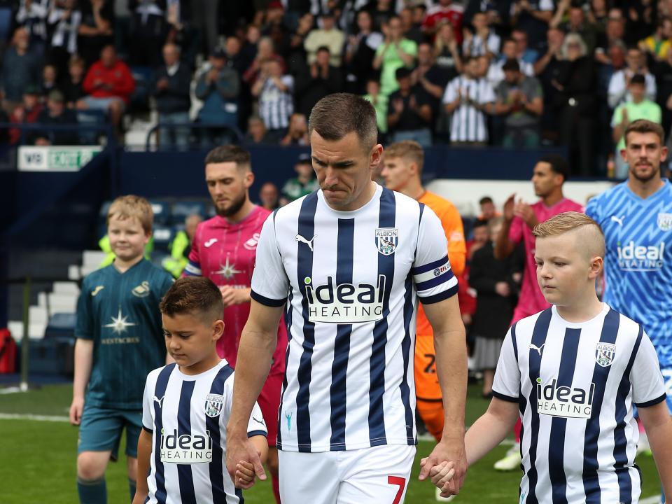 Jed Wallace walking out of the tunnel at The Hawthorns in the home kit with the captain's armband on with two Baggies mascots 