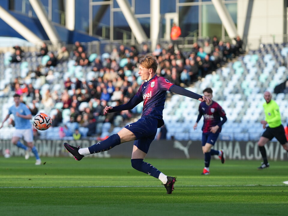 A photo of Ollie Bostock, in the red and blue 23/24 kit, kicking the ball in a PL2 match v Man City