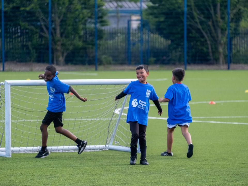Three players wearing blue bibs smile and laugh during the South Asian Emerging Talent Festival on the Academy pitch.