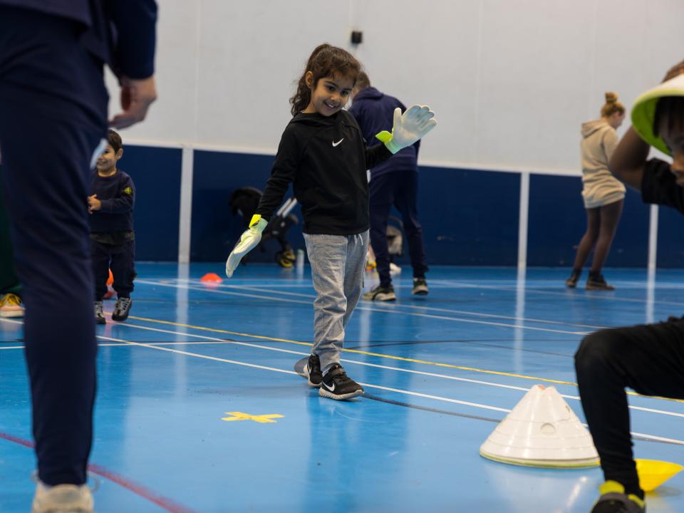 Khadija walks forwards on the blue sports hall surface wearing her goalkeeper gloves.