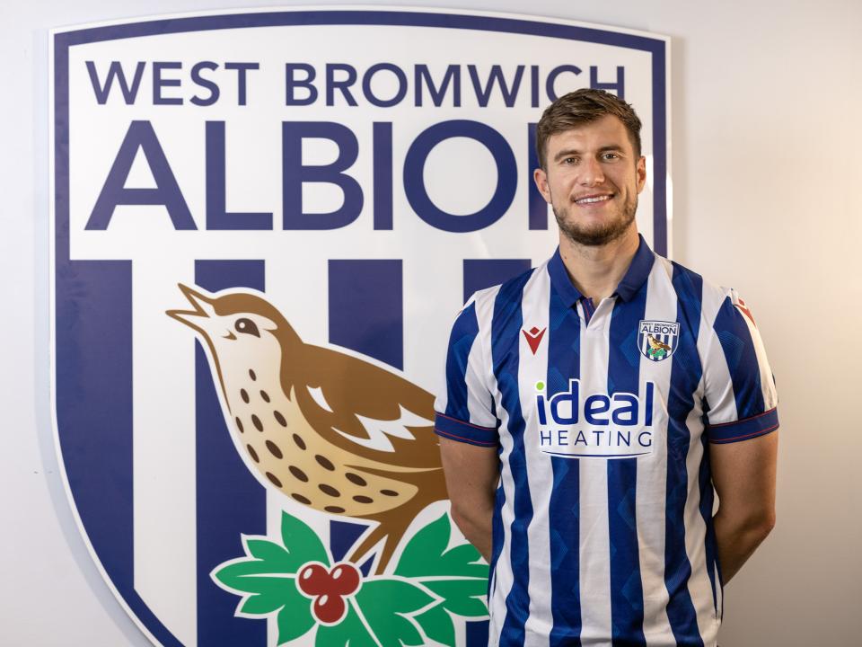 Paddy McNair smiling at the camera while wearing a home shirt stood in front of an Albion badge