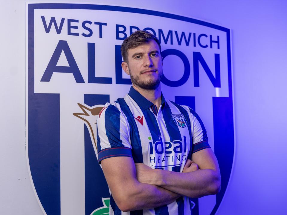 Paddy McNair smiling at the camera while wearing a home shirt stood in front of an Albion badge
