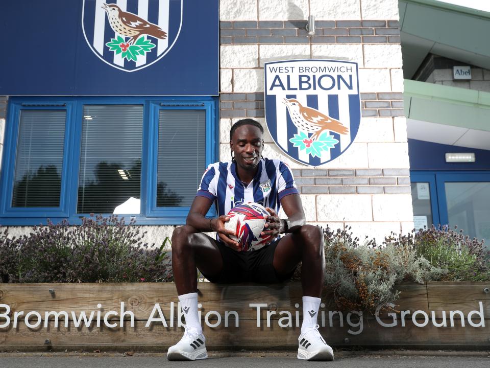 Devante Cole smiling while looking down at a ball sat outside the training ground entrance in a home shirt 