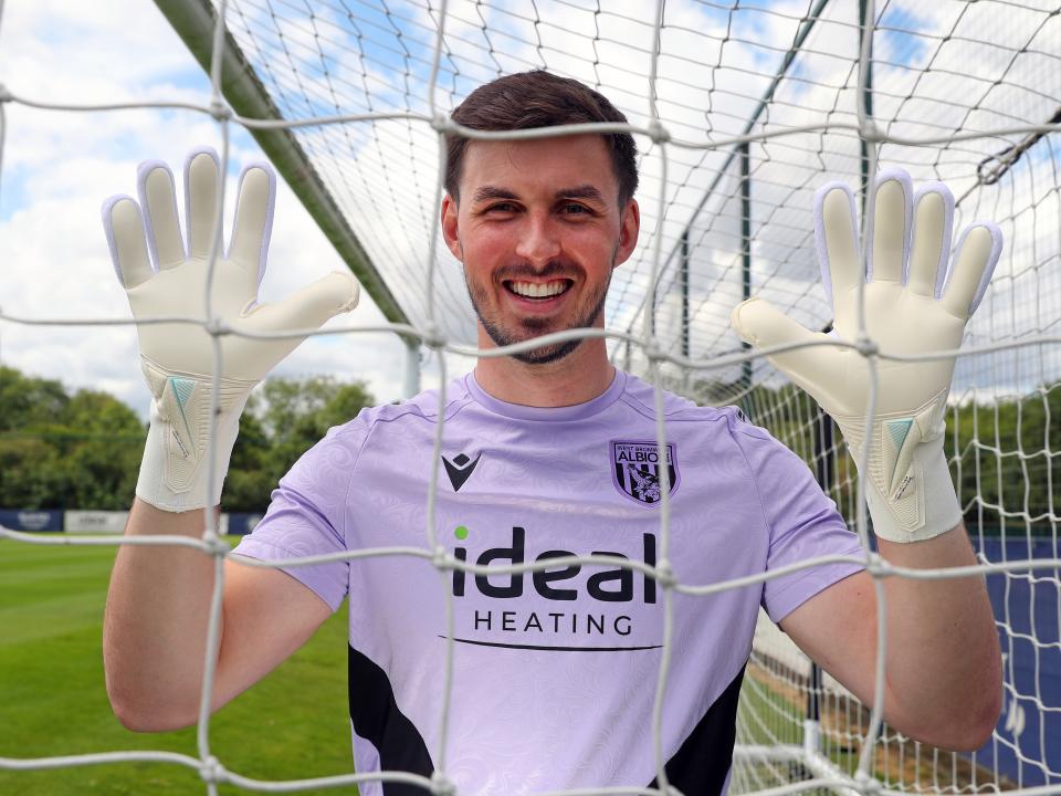 Joe Wildsmith smiling at the camera while holding his gloves up and looking through a goal net