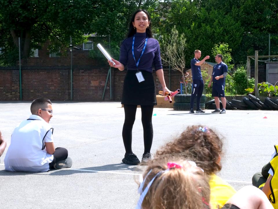 Miss Mendoza addresses the class, as children are seated in lines, in the playground.