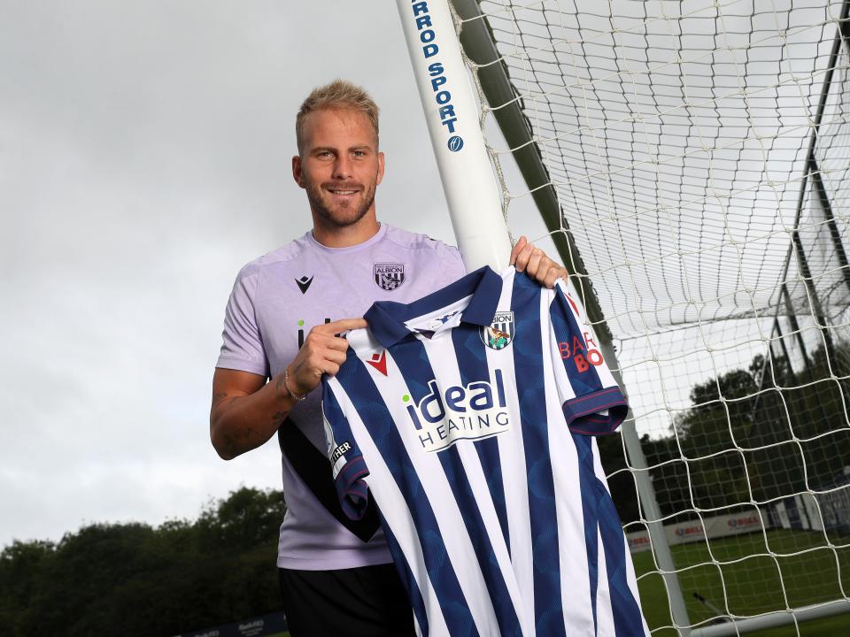 Uroš Račić smiling at the camera while stood holding up a home shirt 