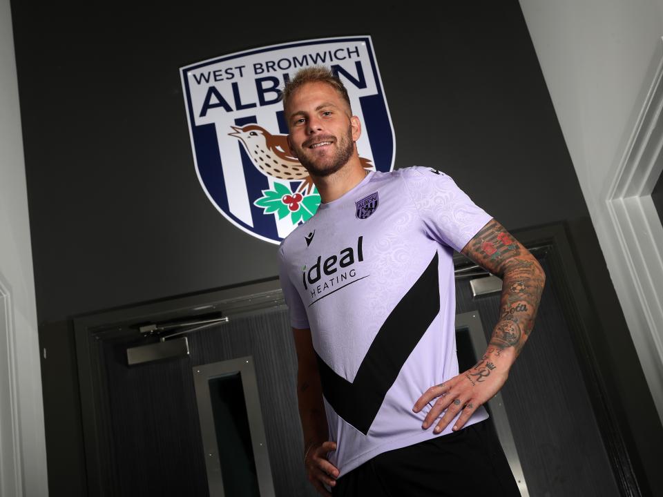 Uroš Račić smiling at the camera while stood in front of an Albion badge 