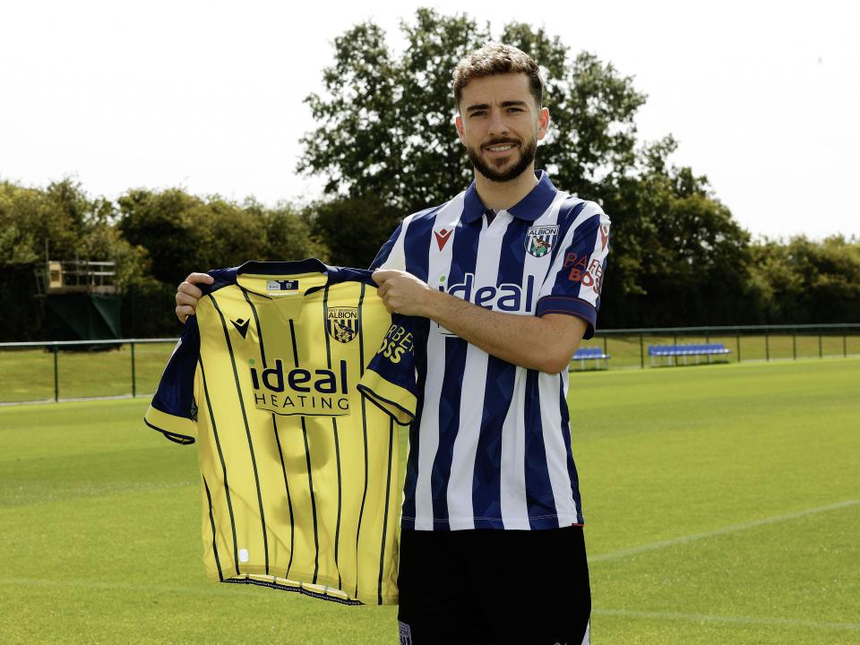 Mikey Johnston smiling at the camera holding up a yellow away shirt while stood in a home shirt 