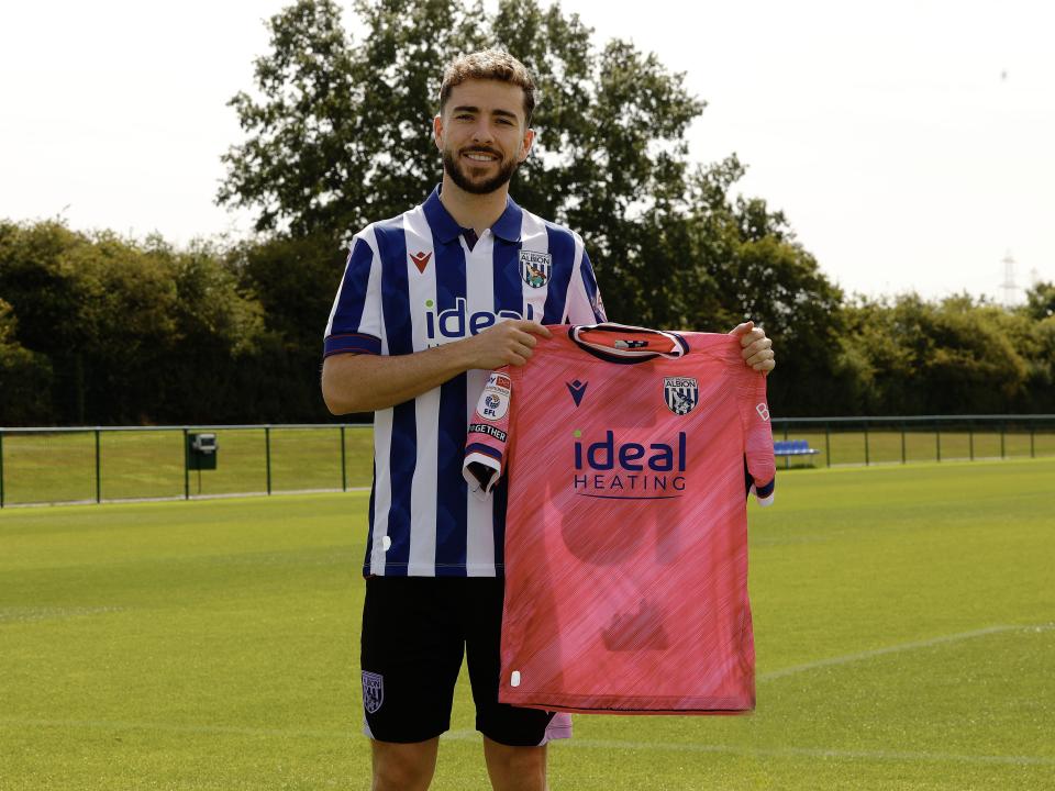 Mikey Johnston smiling at the camera holding up a pink away shirt while stood in a home shirt 