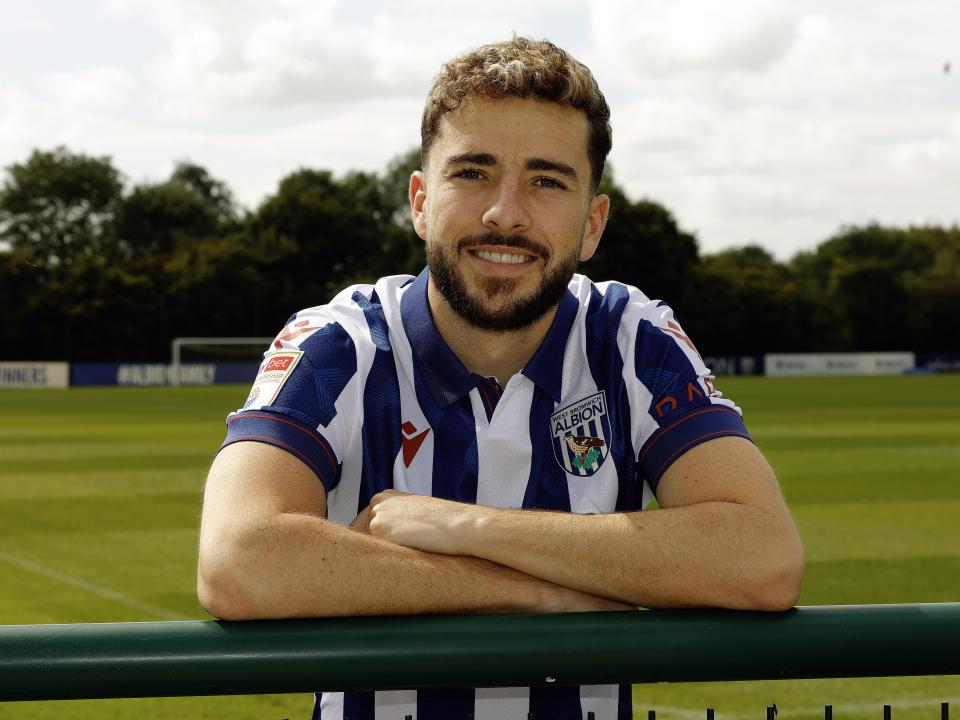 Mikey Johnston leaning over a barrier with his arms folded smiling at the camera while wearing a home shirt 