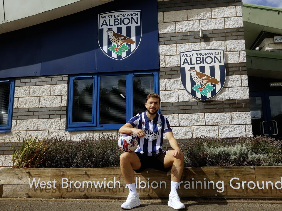 Mikey Johnston smiling at the camera while sat outside the training ground wearing a home shirt and wearing a ball