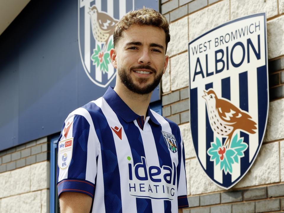Mikey Johnston smiling at the camera while stood outside the training ground in a home shirt 