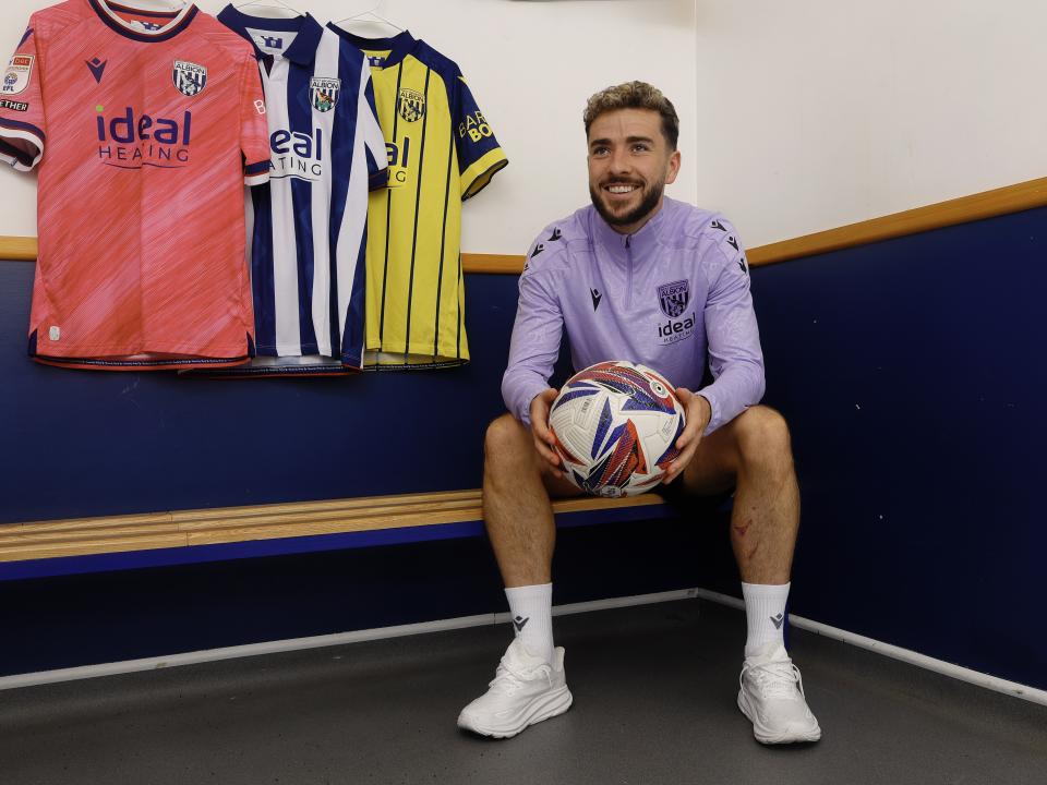 Mikey Johnston sat in a dressing room holding a ball and smiling with all three shirts hanging up behind him