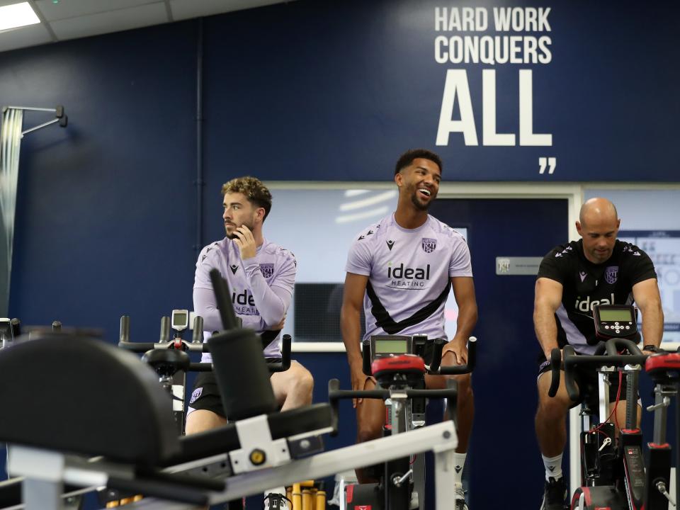 Mason Holgate on a bike in the gym sat next to Mikey Johnston and a member of staff with a smile on his face