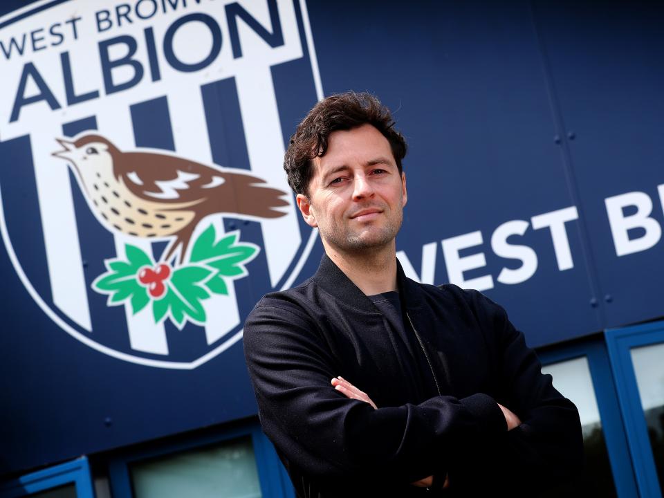 Ryan Mason posing for a photo with his arms crossed and a smile on his face looking at the camera while stood in front of WBA branding 