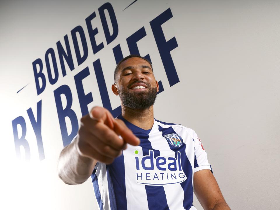 George Campbell smiling and pointing at the camera while sat in a WBA home shirt in front of branding which reads 'bonded by belief'