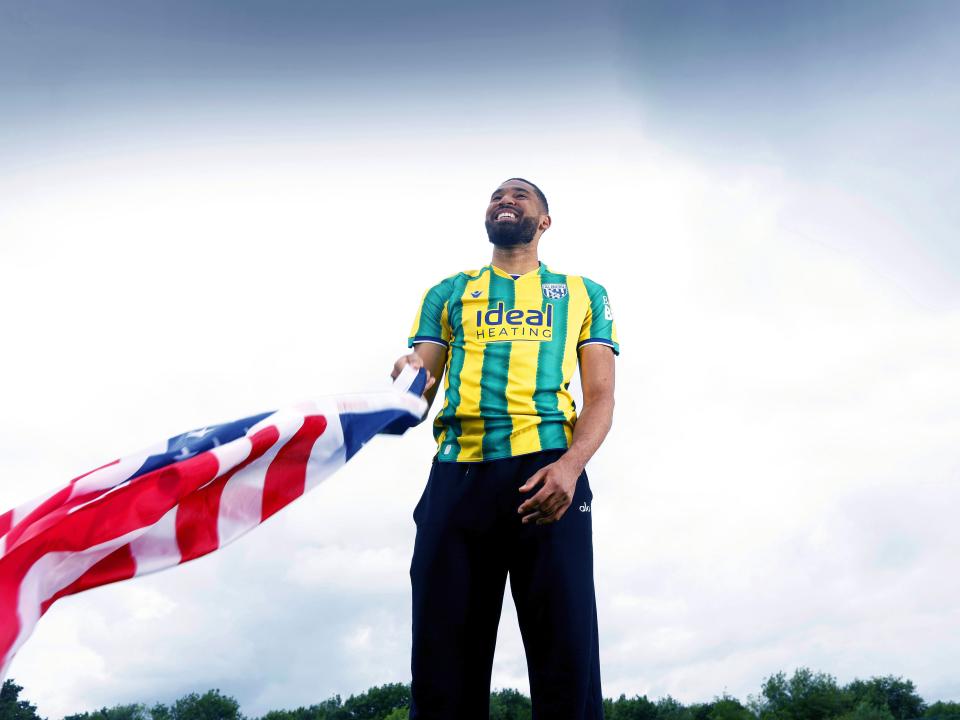 George Campbell smiling while stood in an away WBA shirt waving an American flag