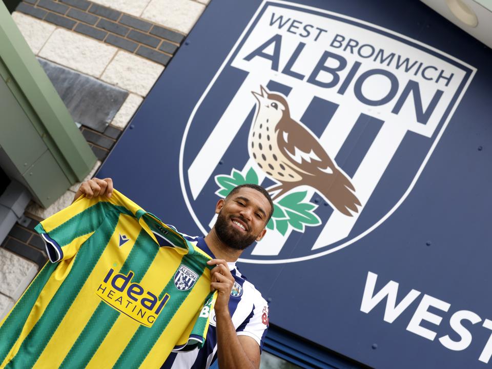 George Campbell holding up an away WBA shirt and smiling at the camera while stood in front of WBA branding