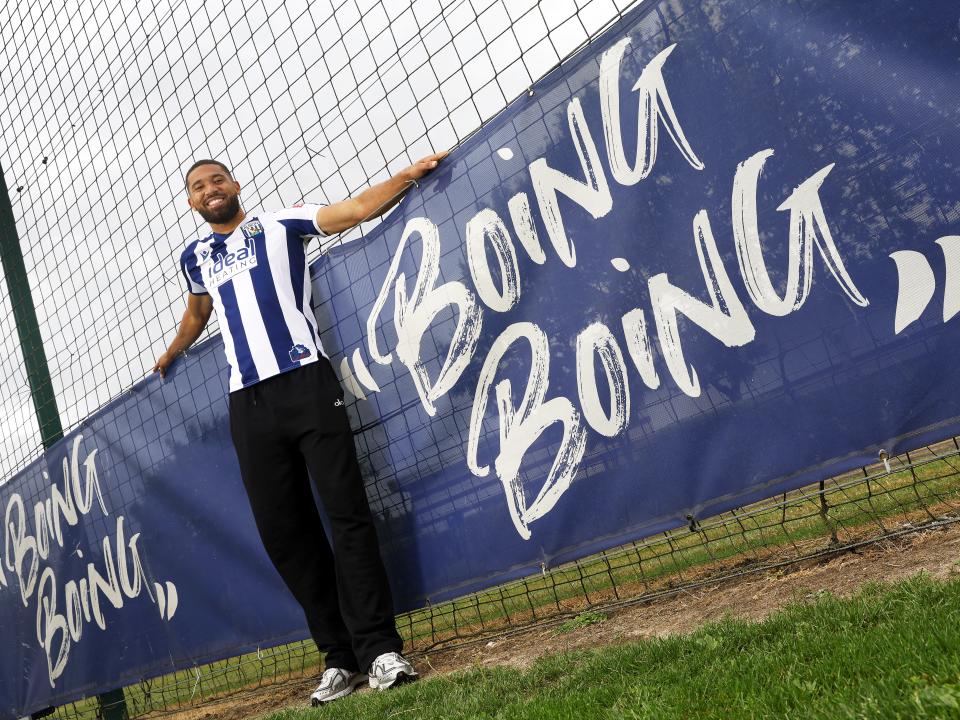 George Campbell stood smiling at the camera in a home WBA shirt next to branding on the pitch which reads 'boing boing'