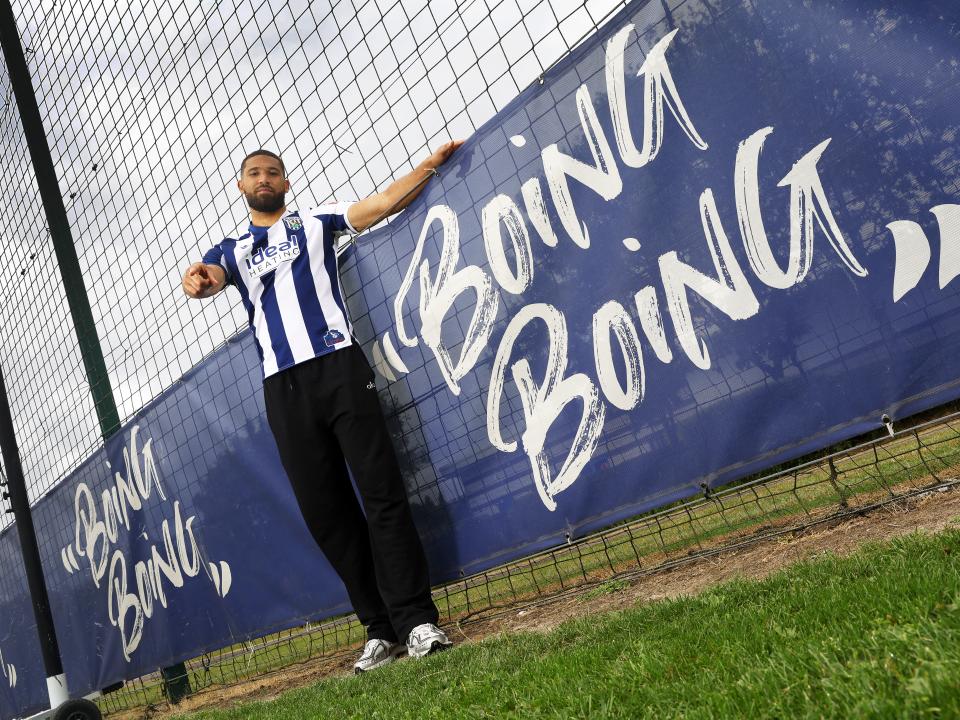 George Campbell stood smiling and pointing at the camera in a home WBA shirt next to branding on the pitch which reads 'boing boing'