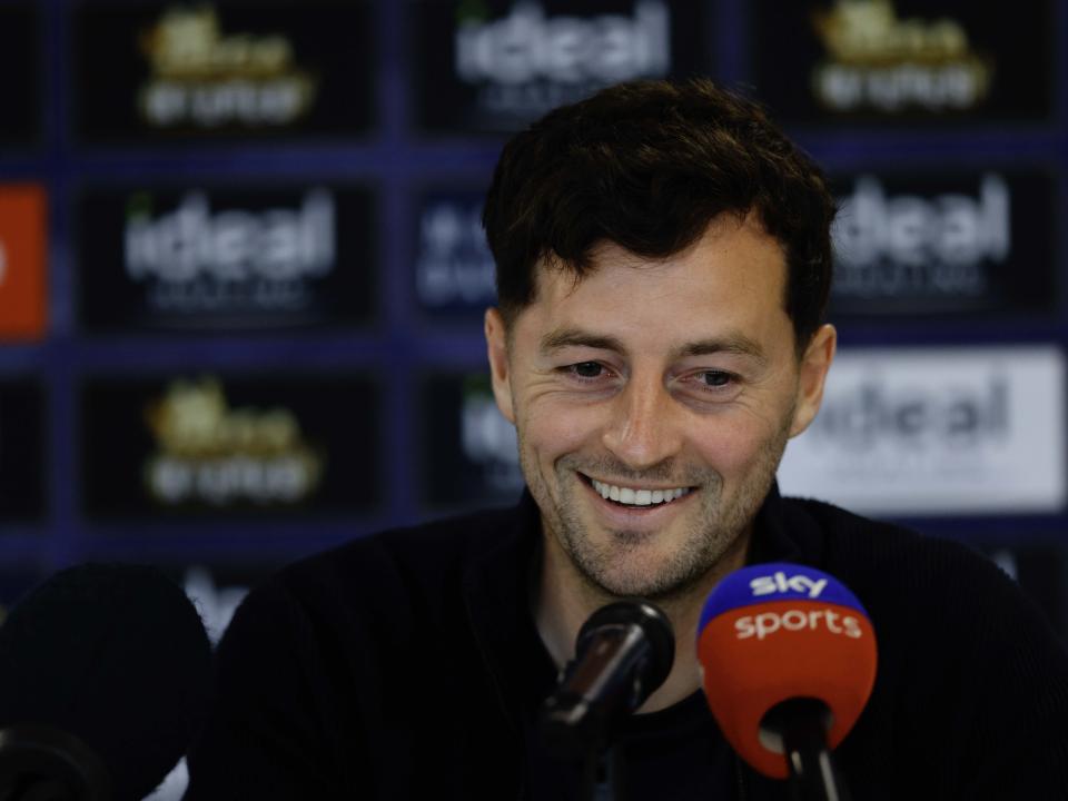 Ryan Mason smiling while talking to the media in front of a sponsor board and behind microphones