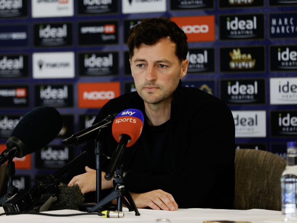 Ryan Mason talking to members of the media in front of a press conference sponsor board and behind a number of microphones