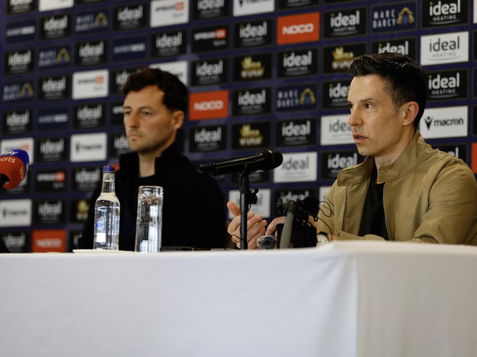 Ryan Mason and Andrew Nestor talking to members of the media in front of a press conference sponsor board and behind a number of microphones