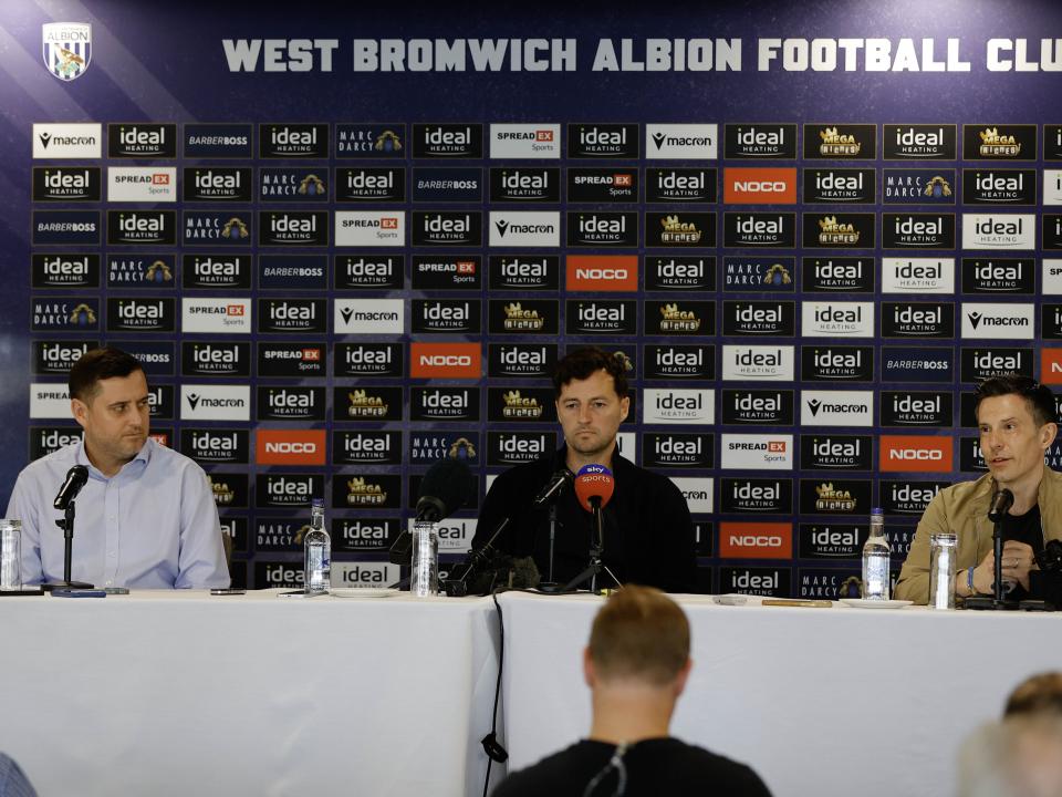 Ryan Mason and Andrew Nestor talking to members of the media in front of a press conference sponsor board and behind a number of microphones