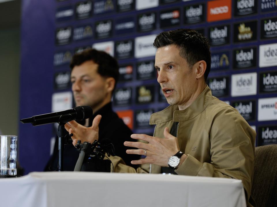 Ryan Mason and Andrew Nestor talking to members of the media in front of a press conference sponsor board and behind a number of microphones