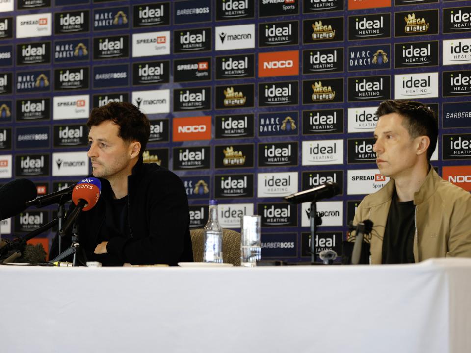Ryan Mason and Andrew Nestor talking to members of the media in front of a press conference sponsor board and behind a number of microphones