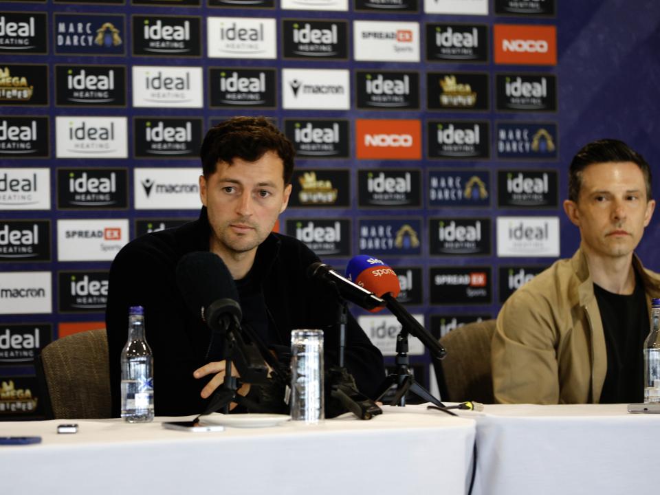Ryan Mason and Andrew Nestor talking to members of the media in front of a press conference sponsor board and behind a number of microphones