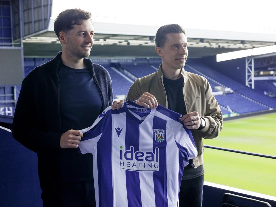 Ryan Mason and Andrew Nestor posing for a photo while holding up a home WBA shirt in front of the West Stand at The Hawthorns