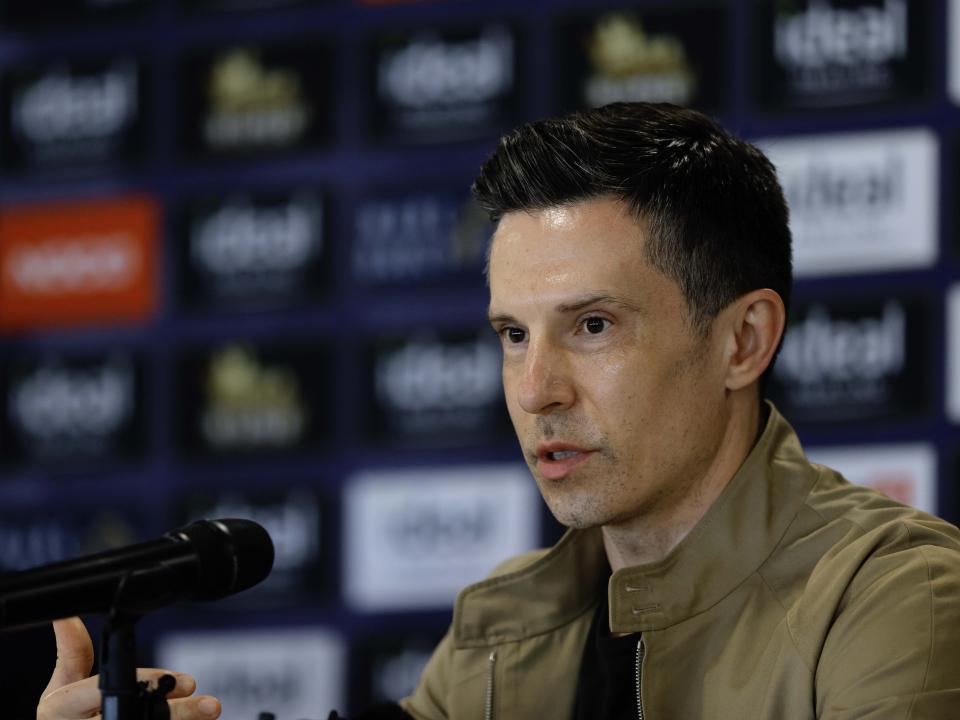 Andrew Nestor talking to members of the media in front of a press conference sponsor board and behind a number of microphones