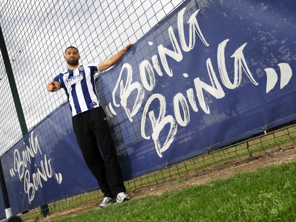 George Campbell smiling at the camera while wearing a home WBA shirt stood next to branding round the training pitch which reads 'boing boing'