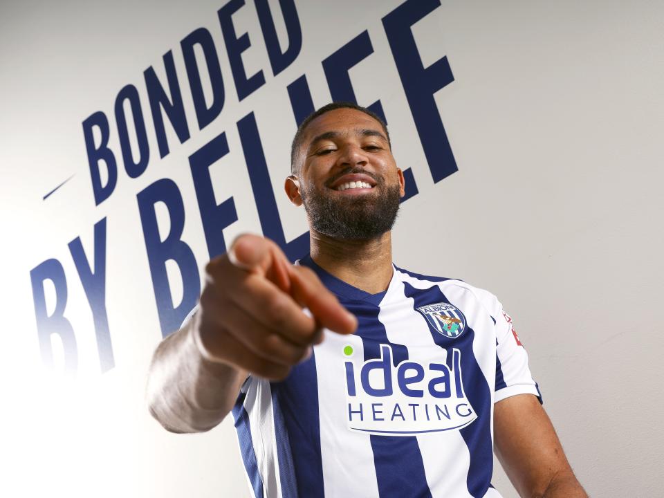 George Campbell smiling and pointing at the camera while in a home WBA shirt with 'bonded by belief' branding written on the wall behind him
