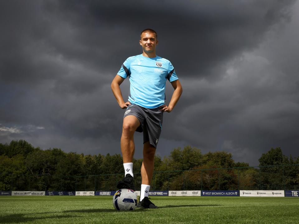 Alfie Gilchrist stood with his foot on a ball and his hands on his hips out on a training pitch smiling at the camera 