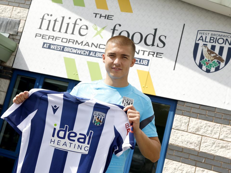 Alfie Gilchrist outside the front of the training ground holding up a home shirt smiling at the camera 