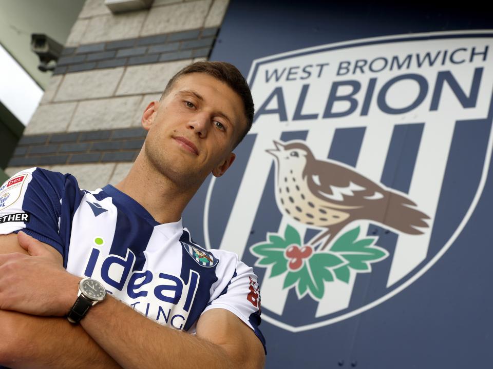 Krystian Bielik with his arms folded in a home shirt stood by a big WBA badge 