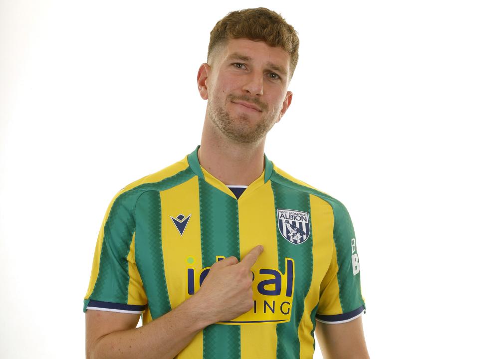 Chris Mepham in the green and yellow away kit smiling at the camera and pointing at the badge