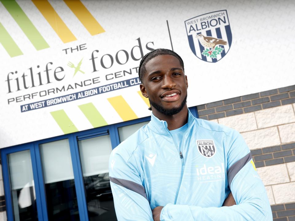 Samuel Iling-Junior smiling at the camera while stood in front of branding outside the training ground 