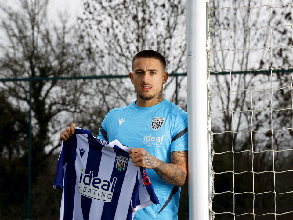 Danny Imray smiling at the camera while holding up a home WBA shirt and leaning against a goal post