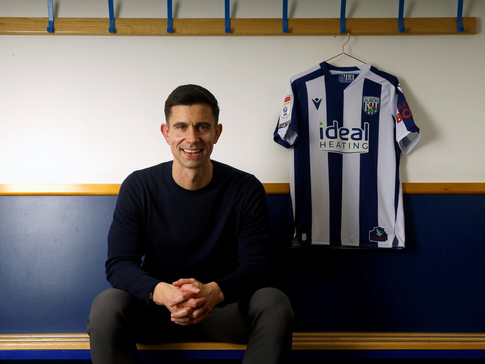 Eric Ramsay sat on a bench in a dressing room smiling at the camera with a home shirt hanging up next to him