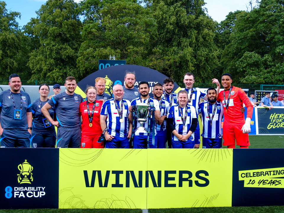 WBA Blind team and coaches with captain Mo Ahmed holding the trophy and EE Disability FA Cup winners banner and arch on display.