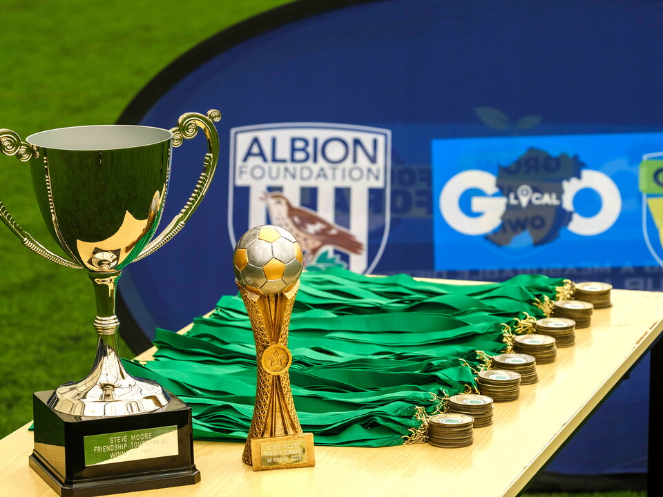 Two trophies and stacked medals on a table with an Albion Foundation branded banner in the background.