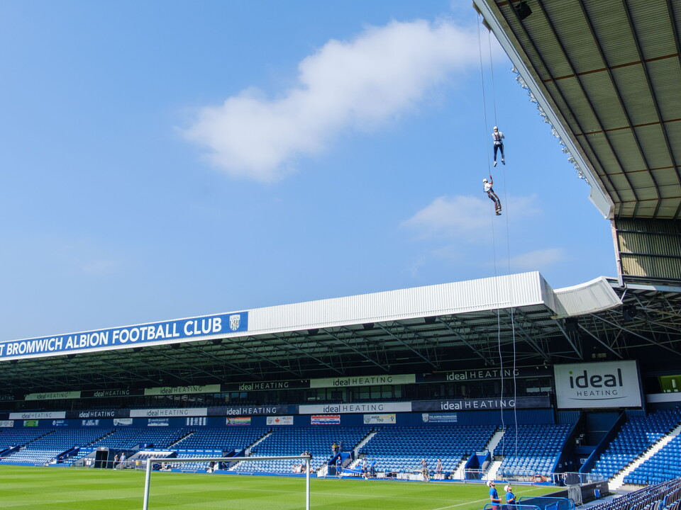 Two participants abseil from the roof of The Hawthorns on ropes suspended high above the pitch, with the West Bromwich Albion Football Club stand and rows of blue seats visible below under a clear sky.