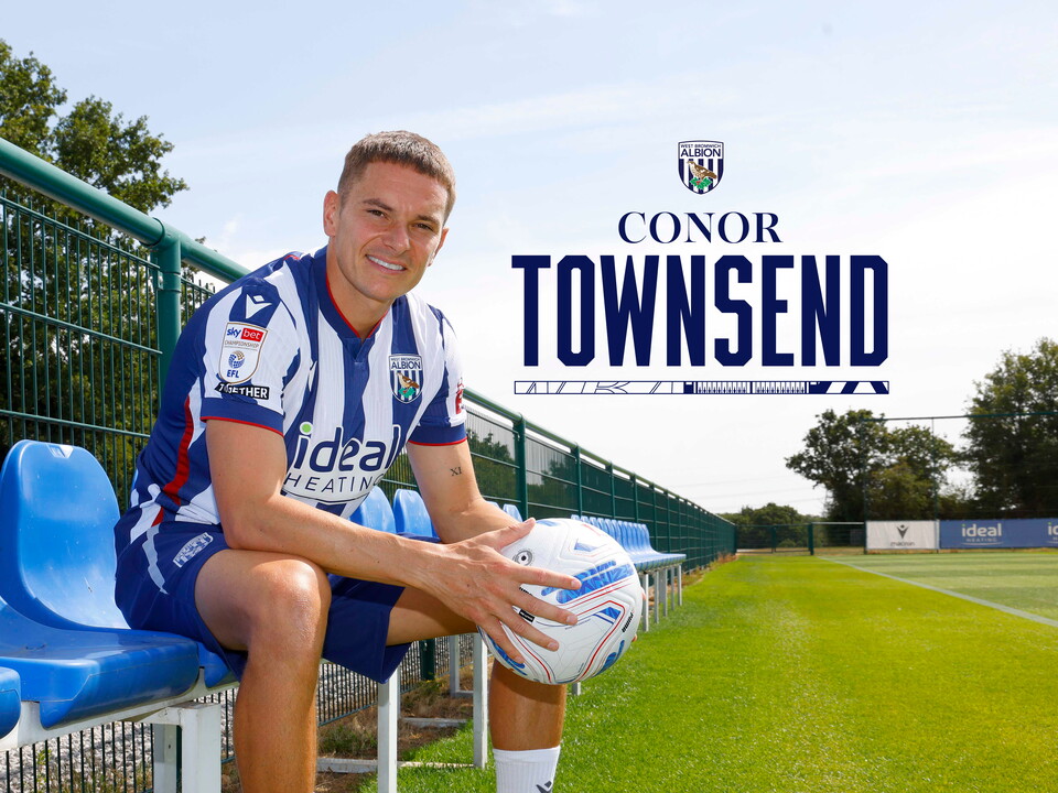Conor Townsend smiling at the camera while sat in a dugout in the home shirt holding a ball