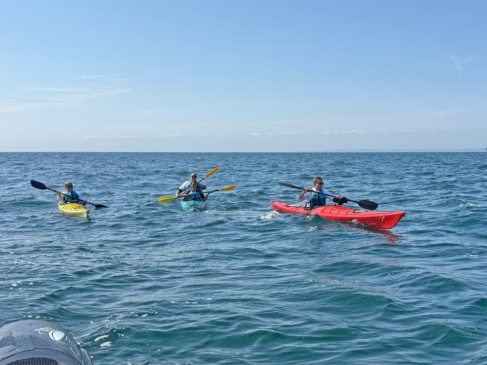 Three people paddle a tandem kayak across open water during an English Channel kayaking challenge, wearing buoyancy aids as they navigate small waves under a clear sky.Provide your feedback on BizChat